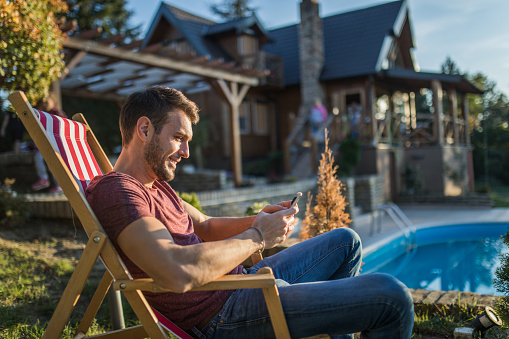 Young man relaxing in deck chair at his back yard and reading a text message on cell phone.