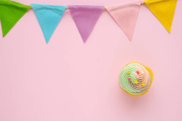 istock Pastel cupcake and colorful bunting party flag on pink background with copy space for text, birthday, anniversary greeting card background, flat lay, top view 956870578