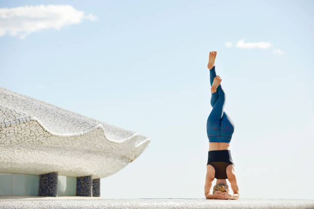 Full length of woman doing headstand Garudasana on building terrace. Flexible female is exercising against sky. She is in sportswear.