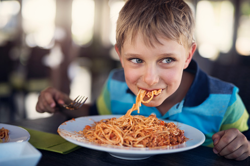 Happy little boy aged 7 is eating spaghetti lunch. The boy is smiling and looking sideways. \nNikon D810