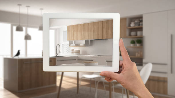 istock Hand holding tablet showing real finished minimalist white and wooden kitchen. Modern blurred kitchen in the background, architecture interior design presentation 956885480