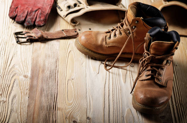 istock Yellow leather used work boots with tool belt and protective gloves on wooden background closeup. Place for text 956895730