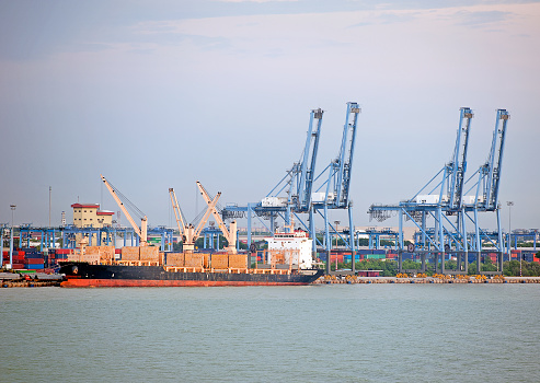 A commercial freighter, a container ship docked alongside the commercial dock by the piers with cranes at the waters edge, manoevering to unload freight, Straits of Malacca, Selangor, Malaysia