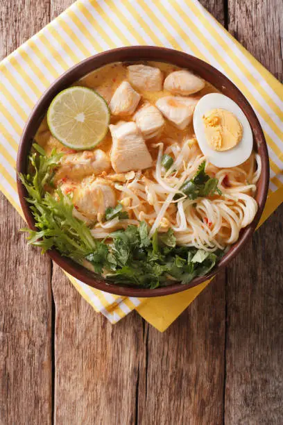 Malaysian laksa soup with chicken, egg, noodles and herbs close up in a bowl on the table. Vertical view from above