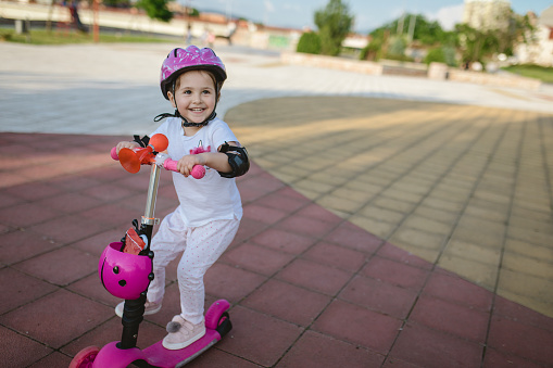 Cute toddler girl wearing a safety helmet and elbow protection while riding a push scooter