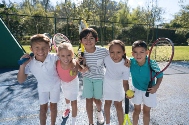 grupo de niños en la cancha de tenis abrazándose uno al otro y sosteniendo sus raquetas mirando a la cámara sonriendo - tennis child childhood sport fotografías e imágenes de stock