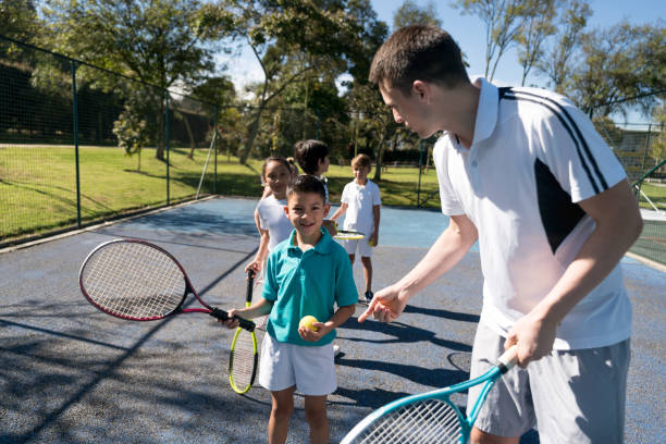 groupe d’enfants en ligne, en attendant de recevoir des instructions de l’enseignant détenant une raquette et une balle de tennis - tennis club photos et images de collection