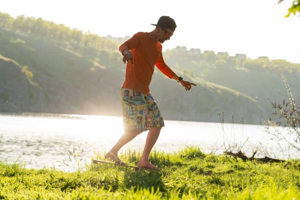 Bearded sporty man is training on the balance board on a green meadow next to river on sunny evening. Back light.