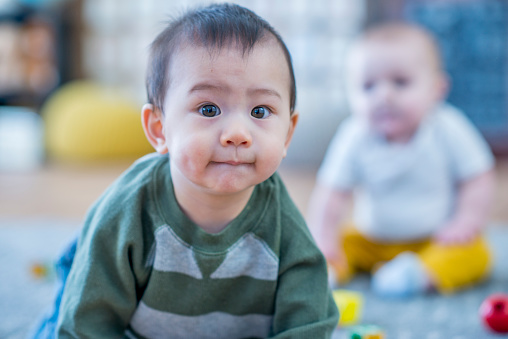 A boy presses his lips together and looks at the camera with big eyes. There is another baby boy playing behind him on the carpet.