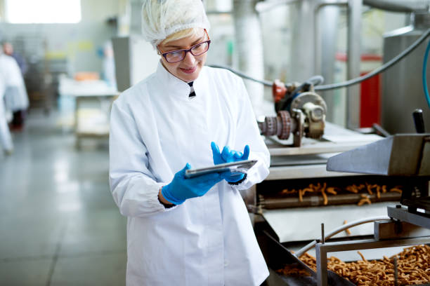 Young cheerful adorable female worker in sterile cloths using a tablet and smiling while standing near the salt sticks factory production line.
