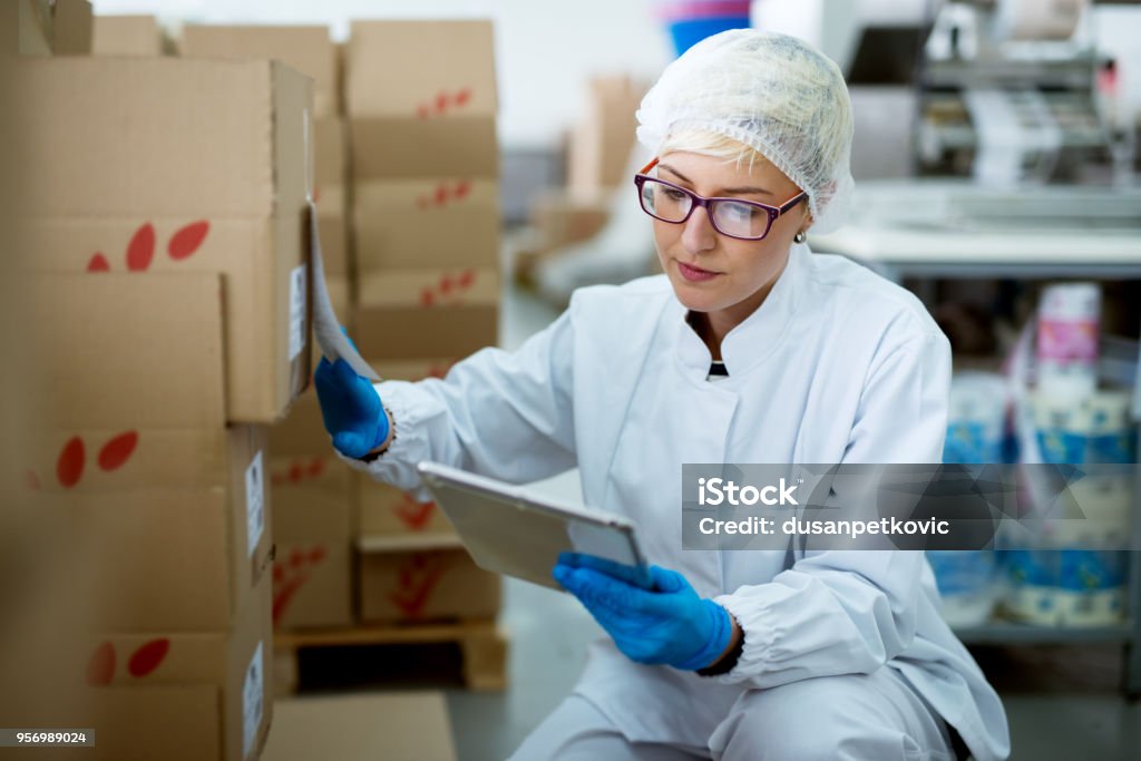 Young beautiful focused female worker in sterile cloths using a tablet to check correction of inventory in factory storage room. Medicine Stock Photo