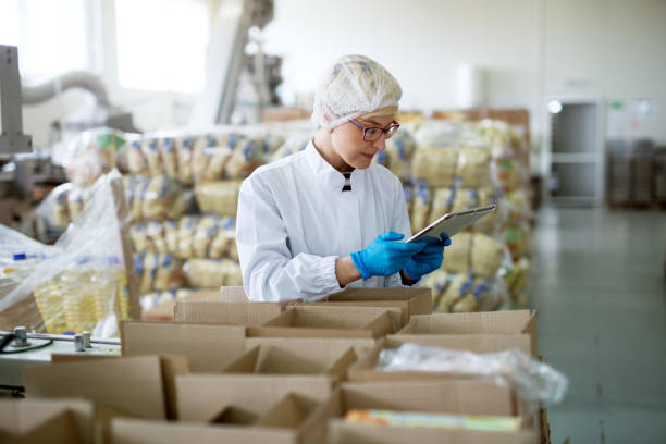 istock Young cute professional female worker in sterile cloths is using a tablet inside a factory storage room while being leaned against boxes. 956989944