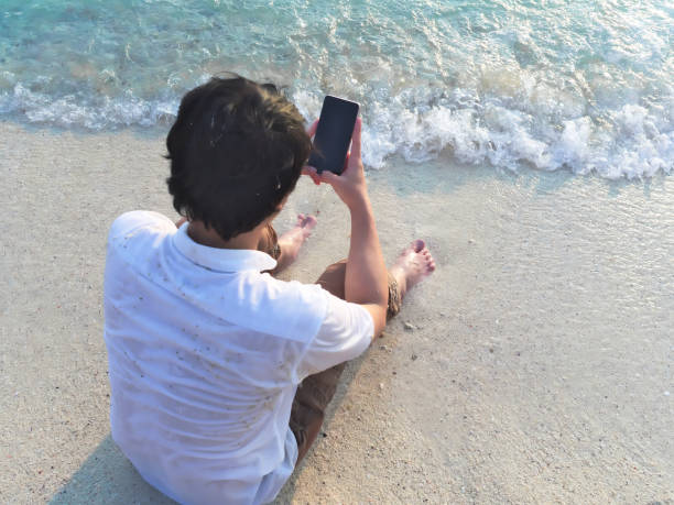 istock Young Asian man using mobile smart phone on the sandy beach. Summer vacations concept. 956990736