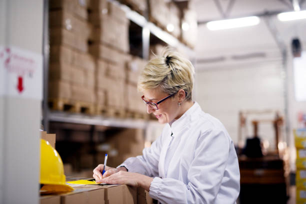 istock Young gorgeous female worker is leaned against the stack of boxes and is writing notes on her yellow folder in factory storage room. 956993704