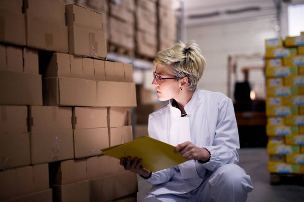 istock Young focused female worker is taking a knee and counting the boxes while holding a yellow folder in factory storage area. 956994390