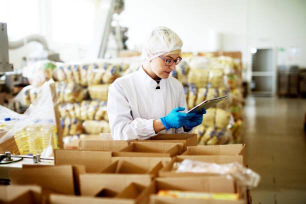 istock Young beautiful worried female worker in sterile clothes is using a tablet while being leaned against the stacks of open boxes inside of factory storage. 956994520