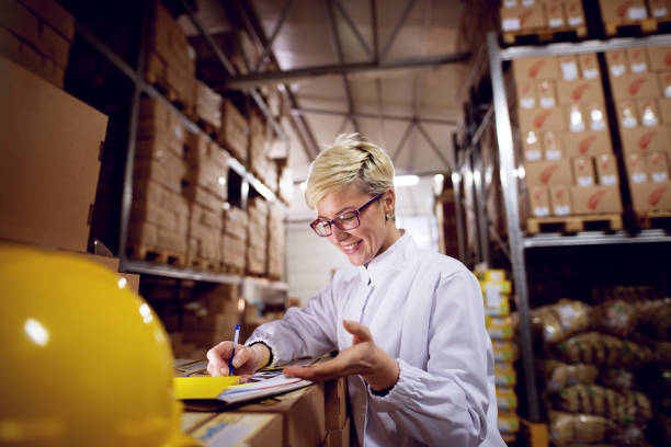 istock Young beautiful cheerful female worker is filling a paperwork on a stack of cardboard boxes while extanding hand in factory storage room. 957001014