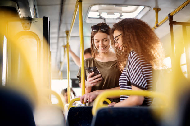 due allegre donne piuttosto giovani sono in piedi su un autobus e guardano il telefono e sorridono mentre aspettano un autobus per portarle a destinazione. - bus transportation indoors people foto e immagini stock