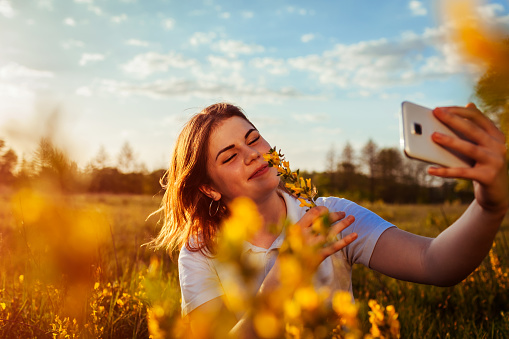 Young woman taking selfie in spring blossoming field at sunset. Allergy free. Happy smiling girl relaxes and enjoys nature.