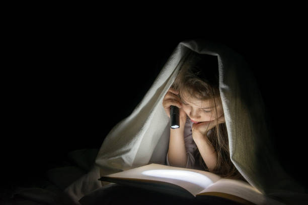 istock Girl with a flashlight under the blanket reading a book in the dark. 957050320