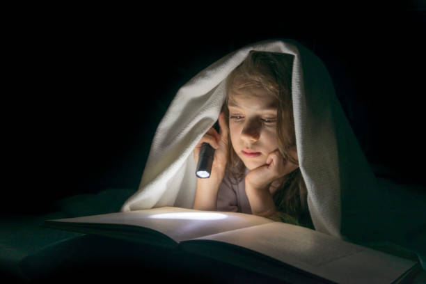 istock Girl with a flashlight under the blanket reading a book in the dark. 957050322