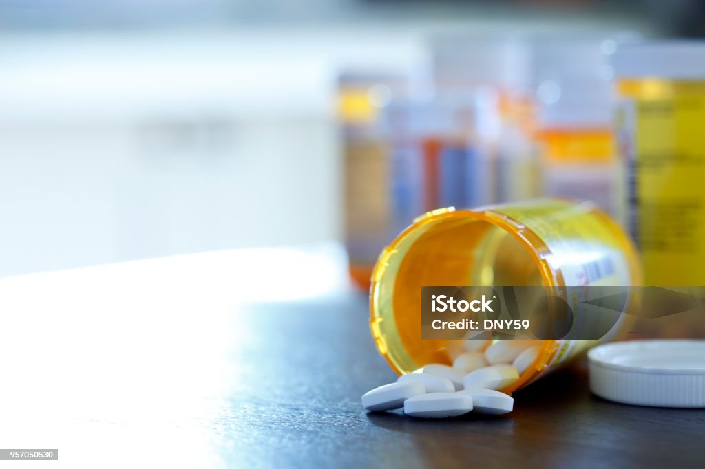 Open Prescription Medication Bottle On Countertop Pills pour out of a prescription medication bottle that lays on its side onto a kitchen counter as a strong morning light filters in through a window. Several other pill bottles stand out of focus in the background. Medicine Stock Photo