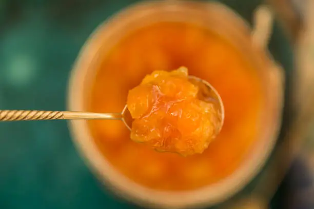 Upper view shot of a tea spoon with orange jam held above a blurred jar with orange jam. Shot with shallow depth of field.
