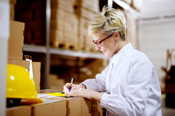 istock Young beautiful happy female worker is filling a paperwork on a stack of cardboard boxes while smiling in factory storage room. 957087466