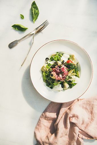 Flat-lay of fresh green summer salad with artichokes, olives, soft cheese and red onion and cutlery in white plate over light marble table background, top view