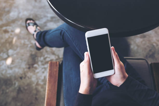istock Top view mockup image of a woman's hand holding white mobile phone with blank desktop screen on thigh in cafe 957116472