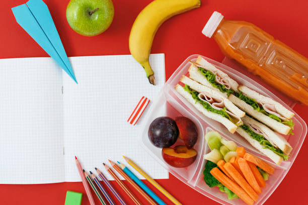 istock Notebook with blank pages with healthy school lunch boxes with sandwich, fresh vegetables, bottle of juice and school supplies on red background. top view. Healthy food concept. Flat lay composition 957125884