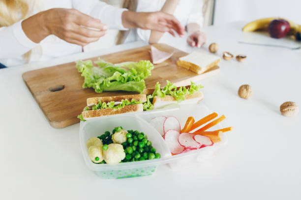 boîtes à lunch école avec sandwich et légumes frais, bouteille d’eau sur la table de cuisine à domicile. dans le fond, la mère et sa fille sont préparent des aliments. concept de la saine alimentation - child human hand sandwich lunch box photos et images de collection