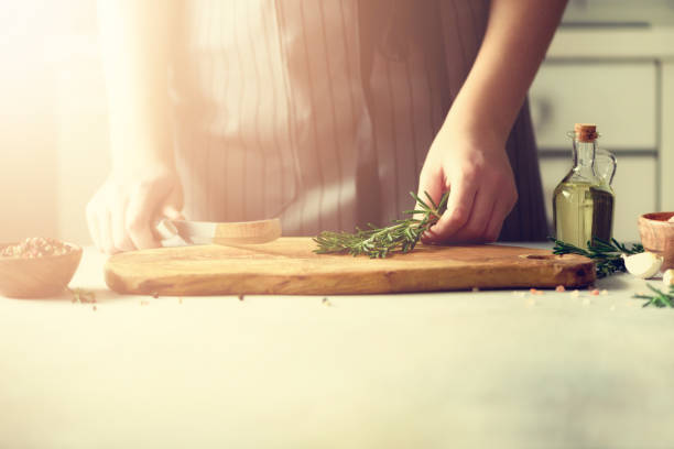 Woman hands cutting fresh green rosemary on wood chopping board in white kitchen, interior. Copy space. Homemade food conceplt, healthy recipe. Take me to work
