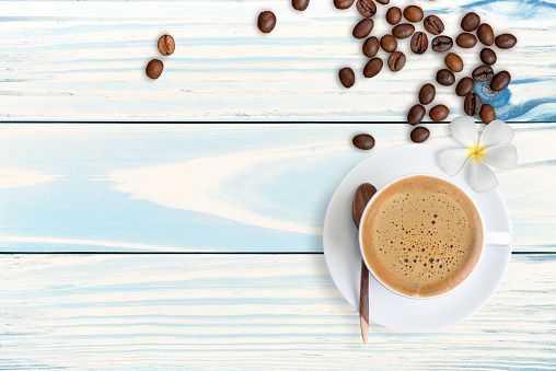 A cup of coffee with plumeria flower and  scattered roasted coffee beans on pastel blue table background, top view.