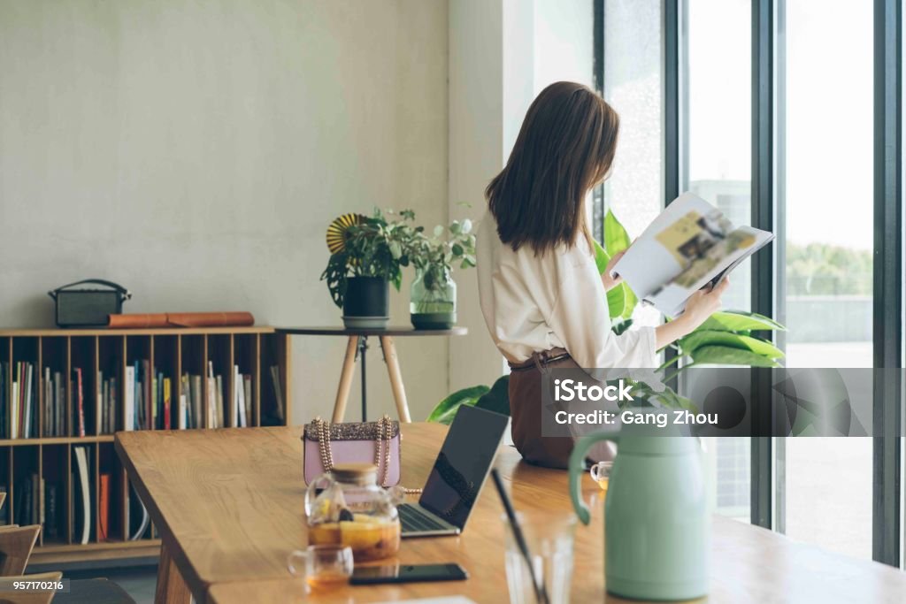 attractive woman enjoying weekend leisure time alone at home attractive woman  reading a fashiong magazine against window at home/cafe. Magazine - Publication Stock Photo