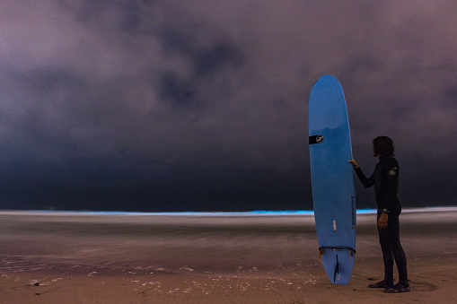 Encinitas, USA - May 9, 2018: Surfer standing on the beach at night after surfing in a bioluminescent algal bloom