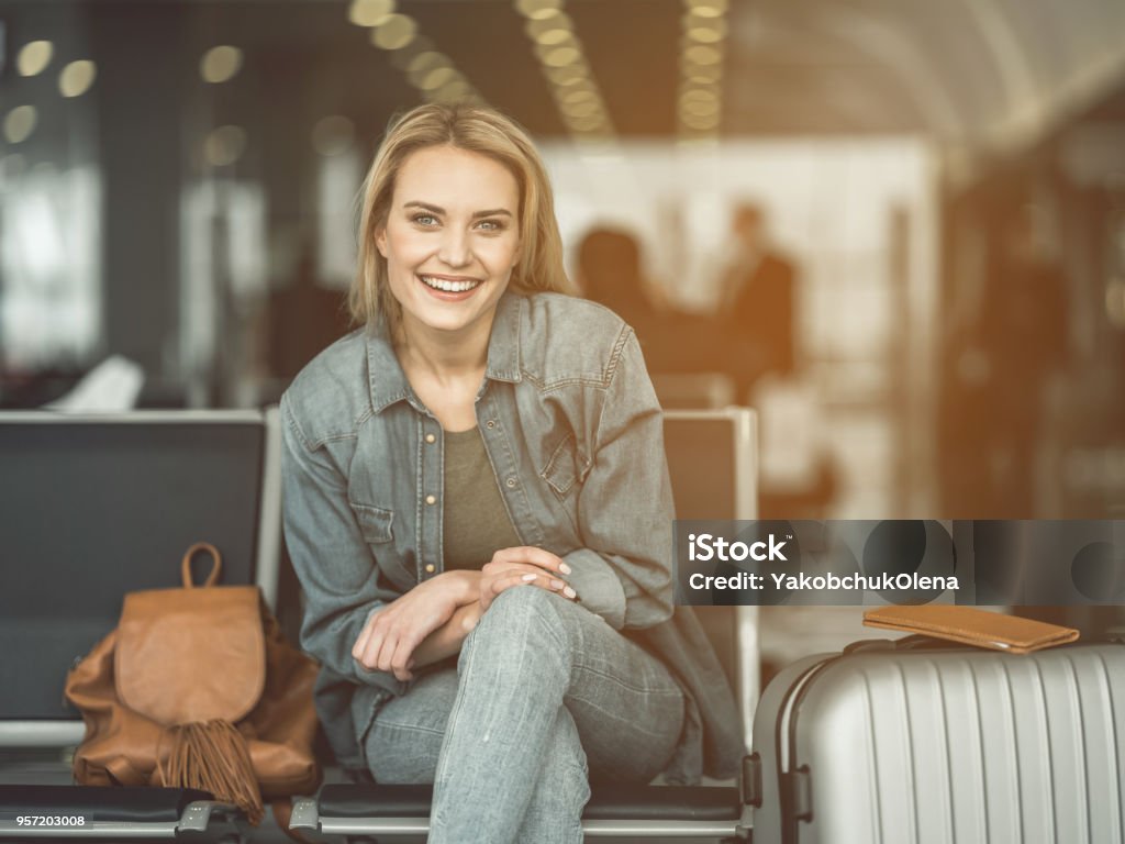 Beaming lady locating in lounge zone Portrait of laughing woman sitting on chair near baggage in airport. Glad tourist waiting for journey concept Airport Stock Photo