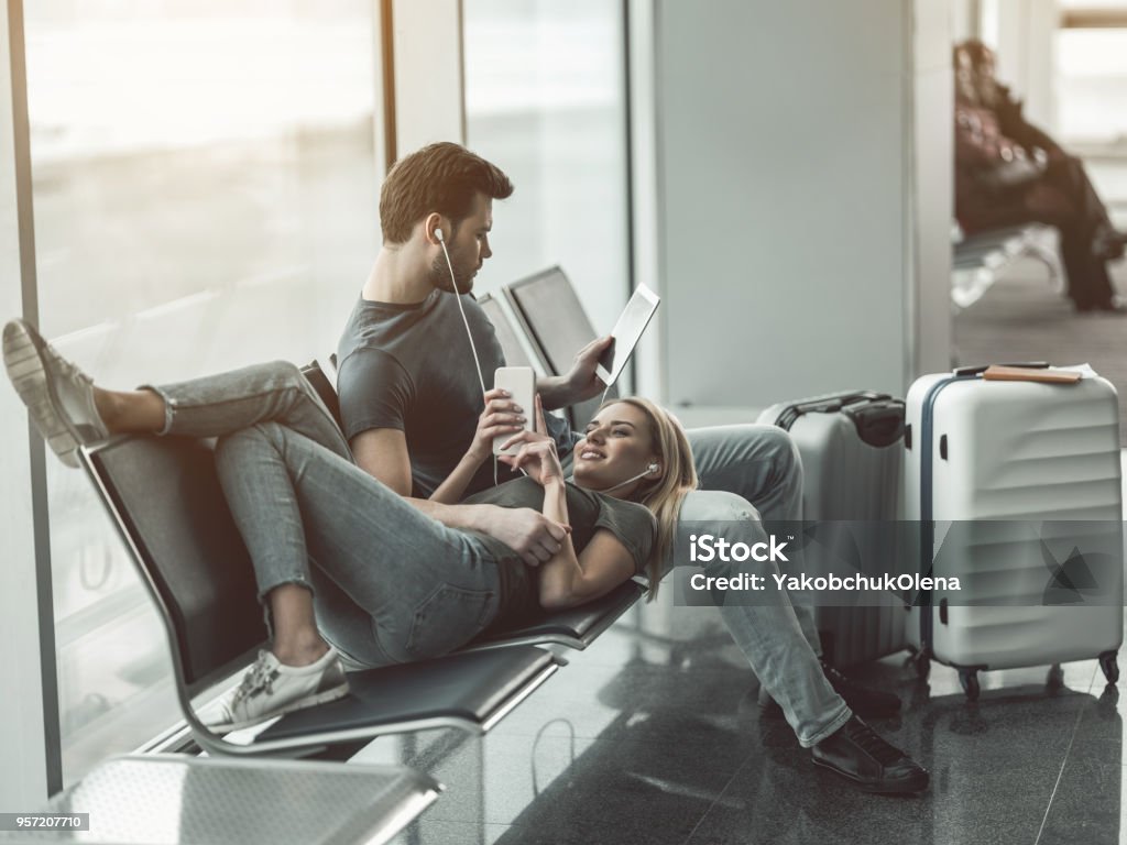 Outgoing lady and boyfriend spending time together I like to spend time with you. Full length beaming woman looking at mobile while lying on male legs. He reading book. Satisfied couple locating in airport concept Airport Stock Photo