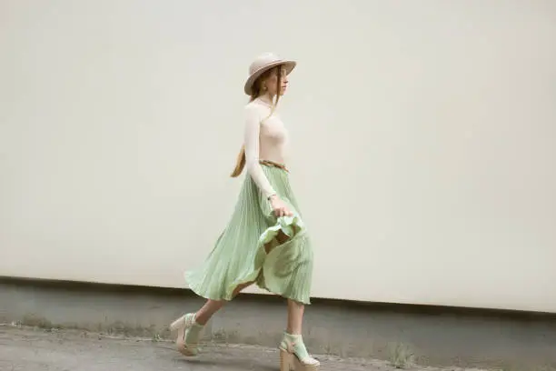 Young red hair girl in hat, dressed in beige blouse and turqoise pleats skirt, on light street backgraund.  Fashion and stylish concept.