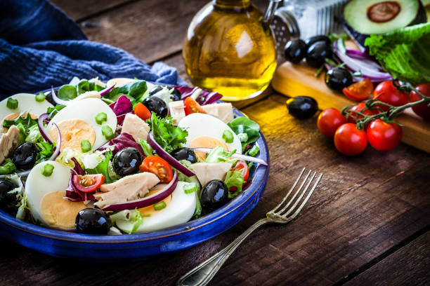 Tuna and hard-boiled eggs salad plate shot on rustic wooden table. Ingredients for preparing salad like lettuce, cherry tomatoes, avocado, olive oil and onion are scattered on the table all around the plate. A table fork is beside the plate. Low key DSRL studio photo taken with Canon EOS 5D Mk II and Canon EF 100mm f/2.8L Macro IS USM