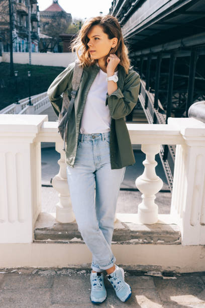 Young woman with wavy hair standing next to a bridge parapet looking aside. Full height photo of fashionable female posing on city street at sunny spring day.