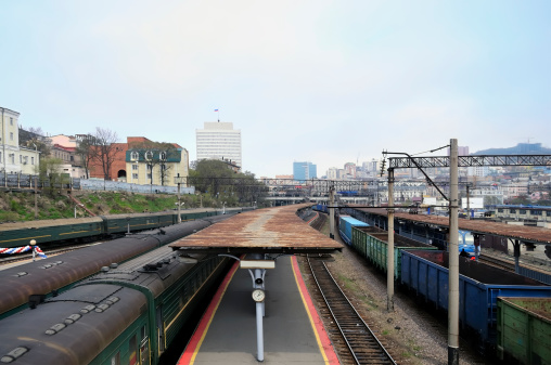 A train station in Vladivostok with passenger and freight cars by the station platform. The badly rusted tin roof vanishes into the city skyline.