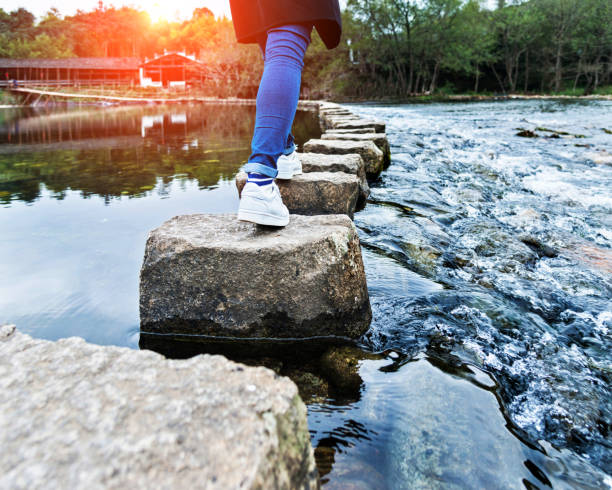 femme traversant les pierres de progression sur une rivière - stepping stone stone stepping footpath photos et images de collection