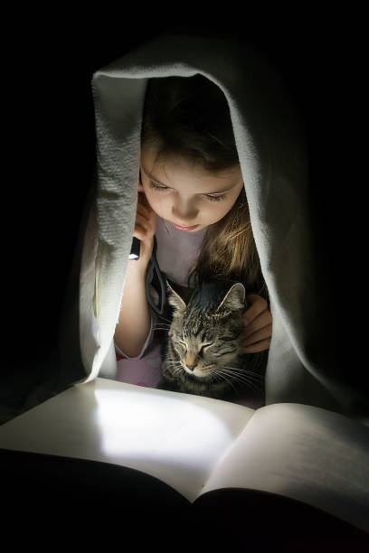 istock Girl with a cat reading a book with a flashlight in the dark under a blanket. 957369238