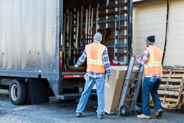 Two multi-ethnic mature workers in their 40s at the back of a truck, loading or unloading a large cardboard box. The men are wearing plaid shirts, reflective vests and jeans. They are moving merchandise for a furniture store.