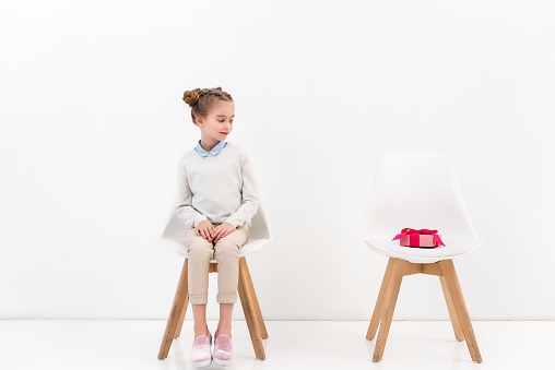 adorable child sitting on chair and looking at present on another chair on white
