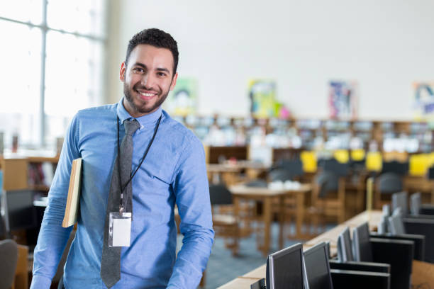 Confident teacher in library Cheerful Hispanic young male teacher stands confidently in the school library and computer lab. He is smiling at the camera. discover card stock pictures, royalty-free photos & images