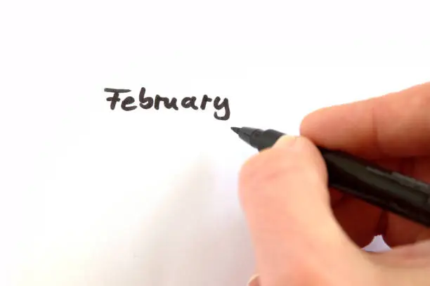February, black handwritten word on white paper, hand holding pen