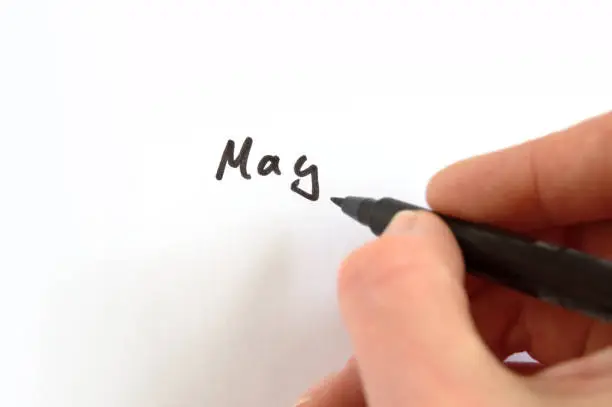 May, black handwritten word on white paper, hand holding pen