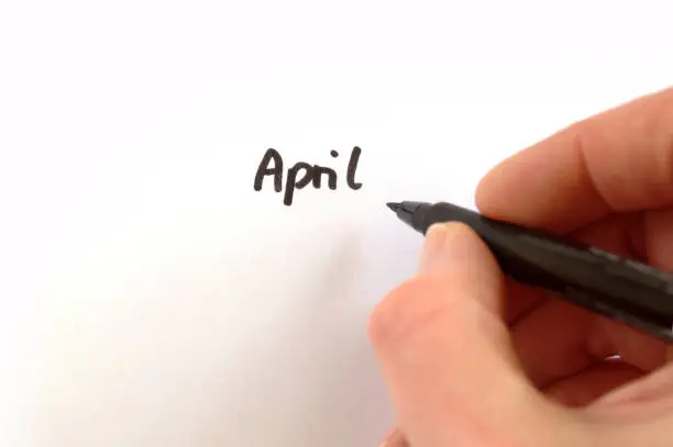 April, black handwritten word on white paper, hand holding pen